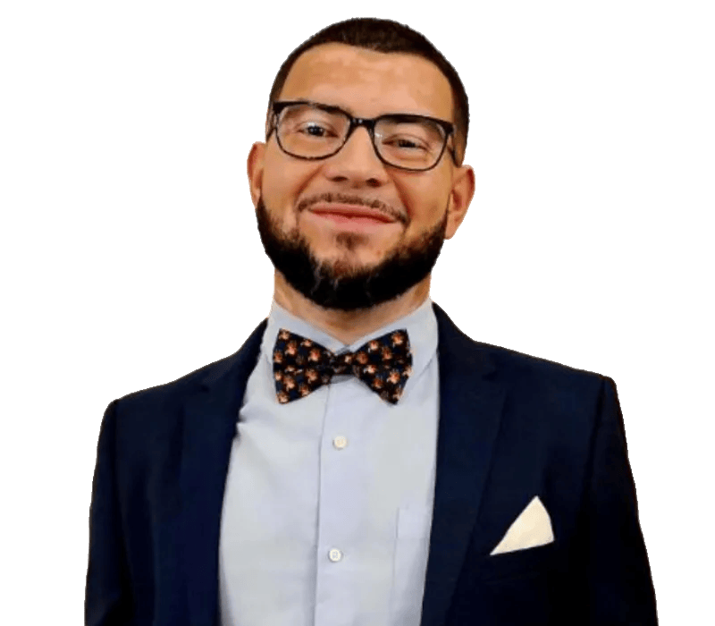 A man with glasses and a beard is wearing a navy suit, light blue shirt, patterned bow tie, and a white pocket square, standing and smiling at the camera.